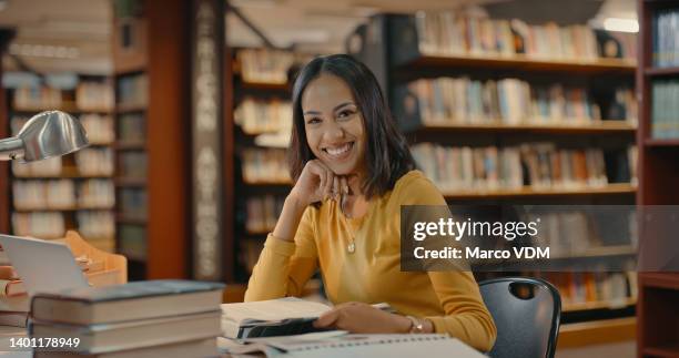 glückliche frau, die allein in einer bibliothek sitzt. junge dame, die an einem projekt arbeitet und in die kamera schaut. gemischtrassige frau, die in einem schreibtisch lächelt und recherchiert - bildtechnik stock-fotos und bilder