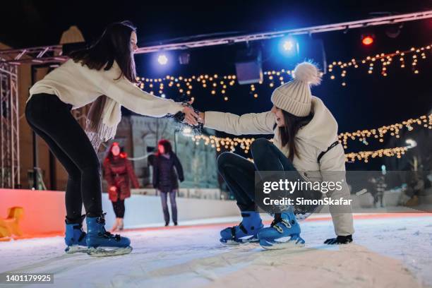 young woman helping female friend to stand up while ice-skating on rink at night - friends skating stock pictures, royalty-free photos & images