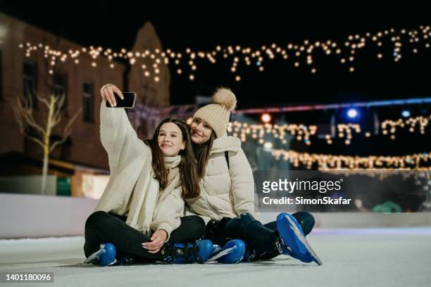 young women taking selfie together with mobile phone while sitting on skate rink at night - ice rink stock pictures, royalty-free photos & images