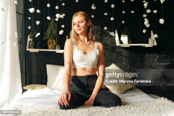 a girl in a white top sits on a large bed by the window. gray background. fluffy blanket. girl with blond hair. room interior. - skinny blonde stock pictures, royalty-free photos & images