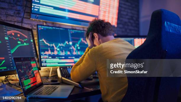 worried man with head in hands sitting in front of stock market graphs on computer screens at desk - distressed stock market people stock pictures, royalty-free photos & images