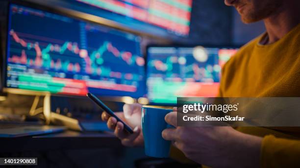man using mobile phone and having coffee while monitoring stock market on computer screens at desk - corretor de bolsa imagens e fotografias de stock