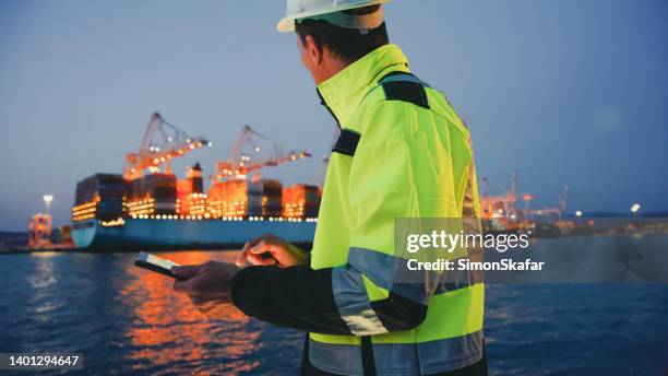 man in protective gear holding tablet computer in front of harbor terminal at night - protective workwear for manual worker stockfoto's en -beelden