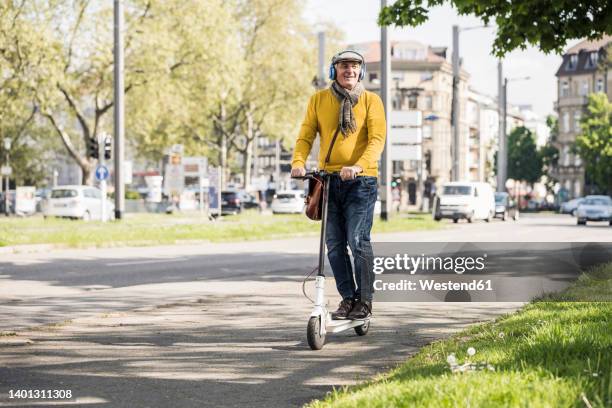 smiling senior man wearing wireless headphones riding on electric push scooter - elderly scooter stock pictures, royalty-free photos & images