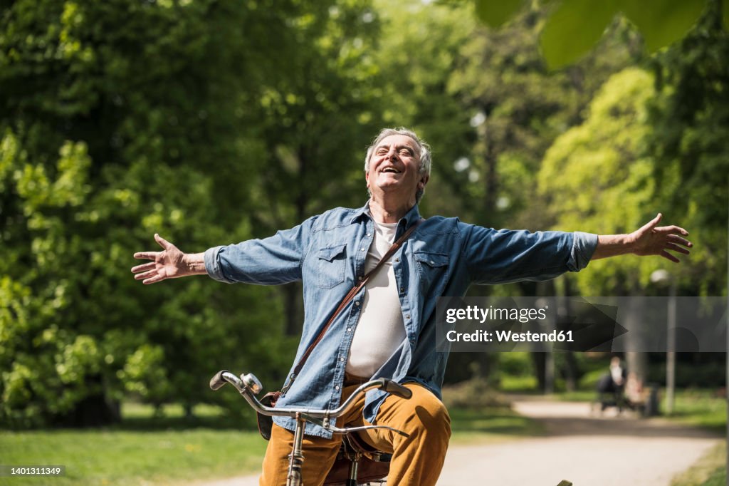 Carefree senior man with arms outstretched riding bicycle at park