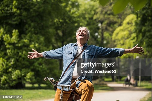 Carefree senior man with arms outstretched riding bicycle at park Carefree senior man with arms outstretched riding bicycle at park