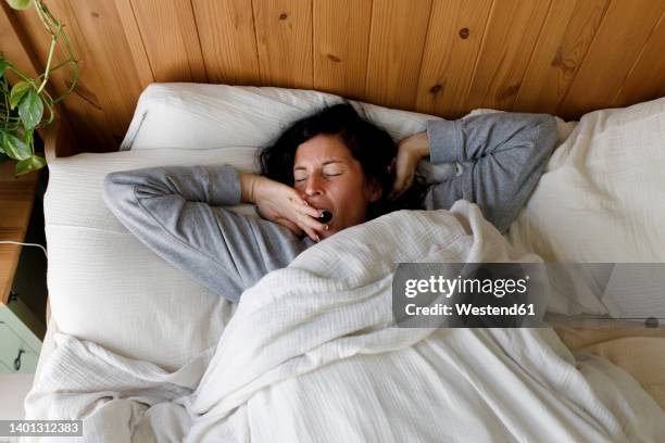 woman with eyes closed yawning in bed at home - waking up bed stock pictures, royalty-free photos & images