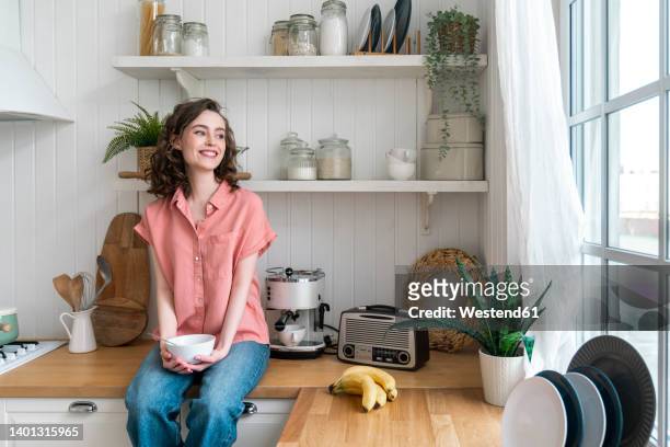 happy woman with bowl sitting on kitchen counter at home - banana woman photos et images de collection
