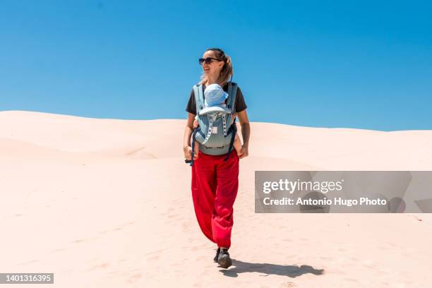 mother carrying her baby across the desert in a baby carrier. - baby carrier stock pictures, royalty-free photos & images