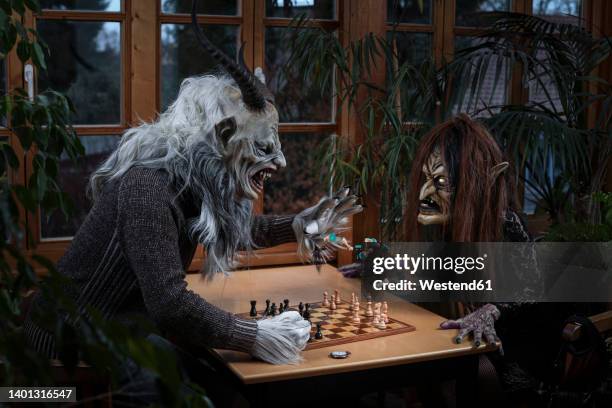 angry man in spooky costume throwing chess piece on witch sitting at table - krampus stock pictures, royalty-free photos & images