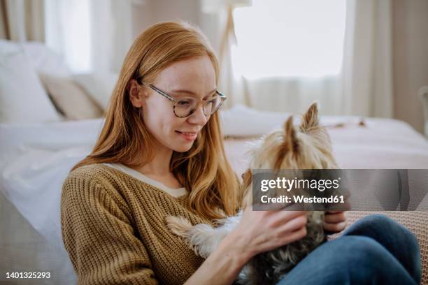 woman wearing eyeglasses sitting with pet dog by bed at home - terrier du yorkshire photos et images de collection