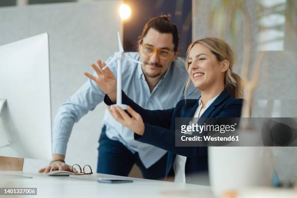 smiling businesswoman holding wind turbine model discussing with colleague in office - sustainability business stock-fotos und bilder