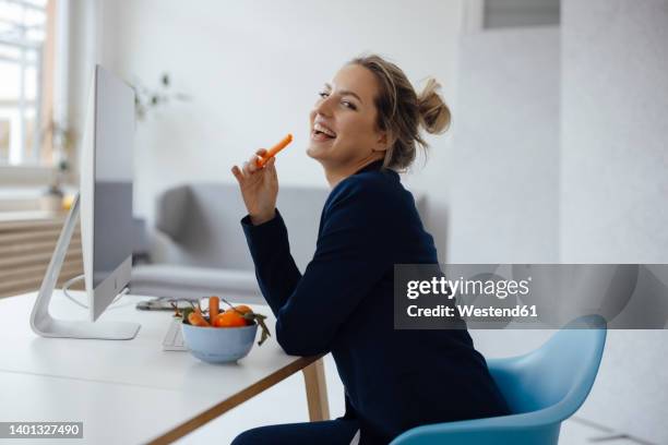 happy businesswoman holding carrot sitting at desk in office - desk with green space view stock pictures, royalty-free photos & images