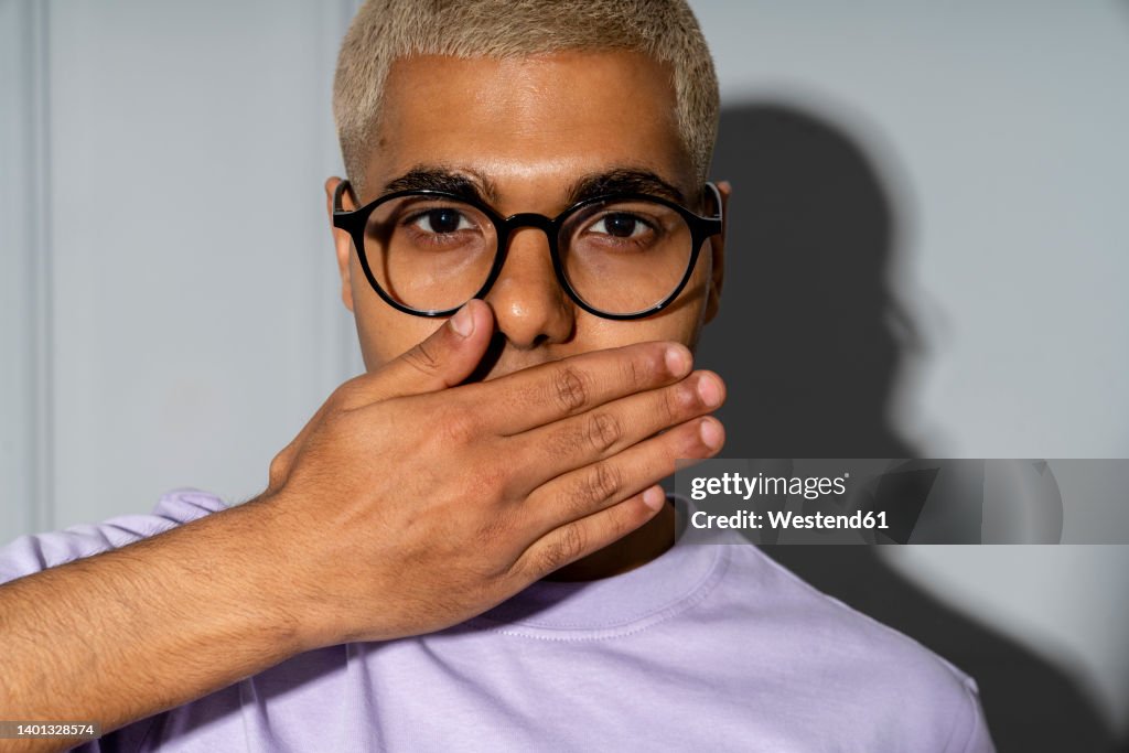 Young man covering mouth with hand in front of wall