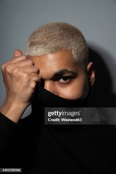 young man covering face with turtleneck top against gray background - man short hair stock pictures, royalty-free photos & images