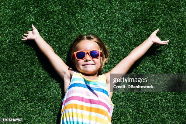 little blonde-haired girl, wearing a colorful summer dress and red-rimmed glasses, lying on the grass sunbathing, arms outstretched, smiling. concept of summer, freedom, relaxation, rest and vacation. - red dress child stockfoto's en -beelden