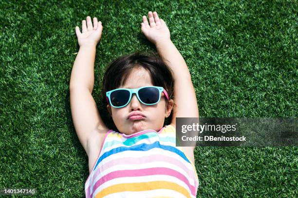 little black-haired girl, wearing a colorful summer dress and red-rimmed glasses, lying on the grass sunbathing, arms outstretched, pouting. concept of summer, freedom, relaxation, rest and vacation. - caprice photos et images de collection