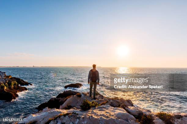 young man standing on a cliff and looking at the sunset by the ocean on baleal island, portugal - portugal coast stock pictures, royalty-free photos & images