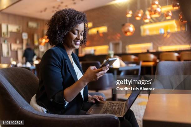 african businesswoman sitting at hotel lounge with laptop using cell phone - african american restaurant texting stockfoto's en -beelden