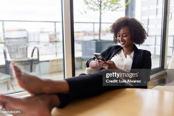 african woman sitting relaxed in hotel room using her mobile phone - african american woman barefoot stock pictures, royalty-free photos & images