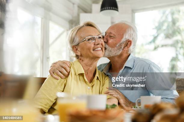 loving senior man kissing his wife during a meal in dining room. - cheek kiss stock pictures, royalty-free photos & images