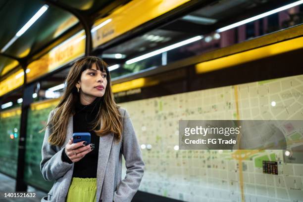 young woman looking away holding a smartphone while waiting for a bus - white night stock pictures, royalty-free photos & images