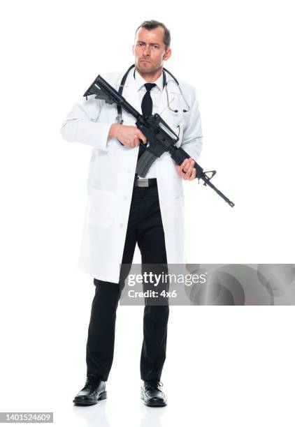 caucasian young male doctor standing in front of white background wearing lab coat and holding weapon and using semi-automatic pistol - machinegeweer stockfoto's en -beelden