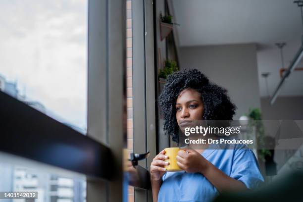 black nurse looking out the window with a cup of coffee in her hand - nurse serious stock pictures, royalty-free photos & images