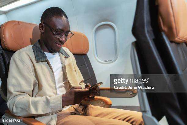 happy black male using a phone texting or playing mobile phone sitting in airplane he's touching smartphone screen - airport business lounge stock pictures, royalty-free photos & images