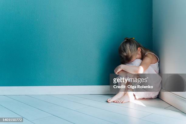 a little sad and depressed blond girl in white dress sitting on the floor in the corner indoors - child abuse stock-fotos und bilder