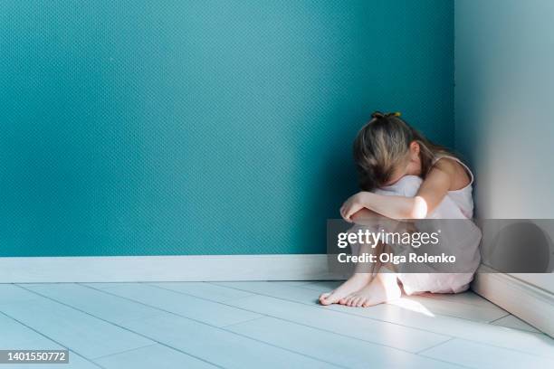 a little sad and depressed blond girl in white dress sitting on the floor in the corner indoors - kindermishandeling stockfoto's en -beelden