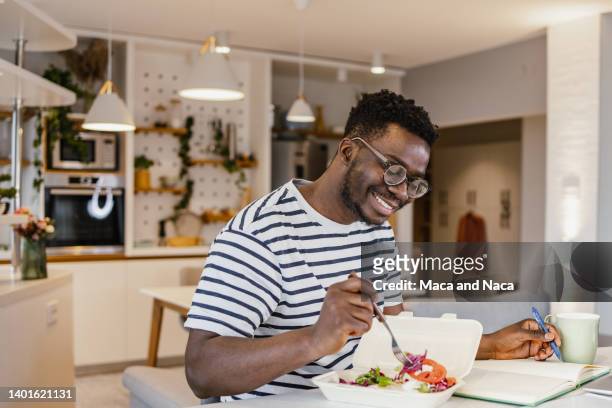 young african-american man eating takeaway meal at home and planning activities - diet journal stockfoto's en -beelden