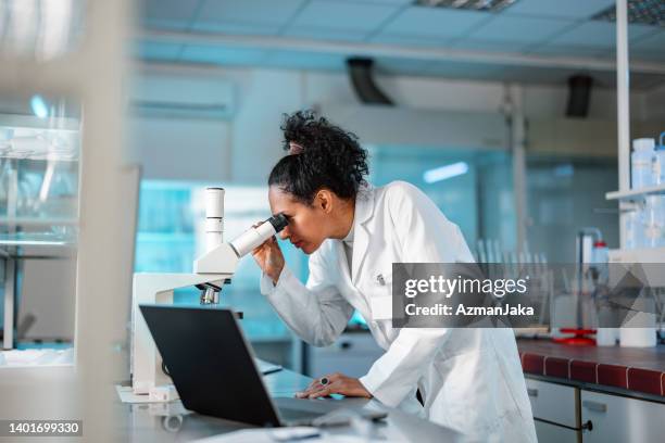 wissenschaftlerin, die unter dem mikroskop schaut und einen laptop in einem labor benutzt - reifen stock-fotos und bilder