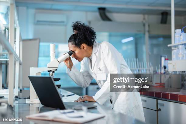 scienziata che guarda al microscopio e usa il laptop in un laboratorio - biochemical foto e immagini stock