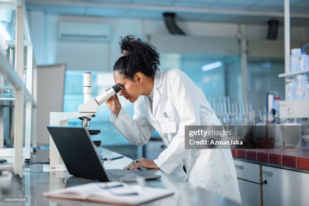 Cientista feminina olhando sob microscópio e usando laptop em um laboratório