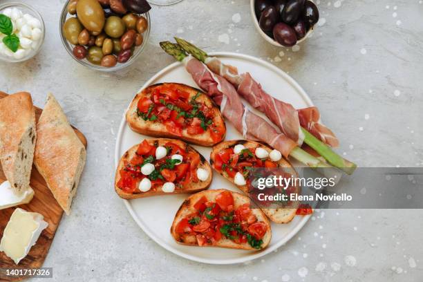 food for lunch and dinner or aperitifs bruschetta with tomatoes and mozzarella, asparagus with bacon.  food antipasto prosciutto ham, salami, olives and bread . cheese on a board parmesan, pecorino, gorgonzola. two glasses of white wine or prosecco - bruschetta stock pictures, royalty-free photos & images