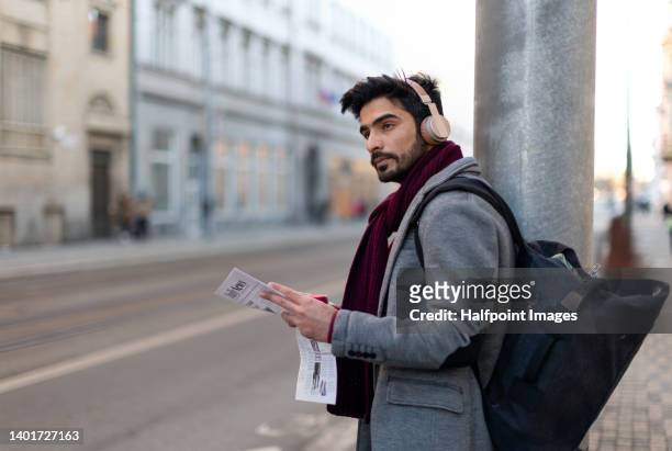 young handsome muslim man commuter waiting on the street, with backpack, headphones, reading newspaper. - handsome muslim men stock pictures, royalty-free photos & images