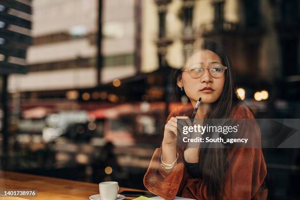 asian woman sitting in the cafe and writing notes while being on the coffee break - gefotografeerd door het raam stockfoto's en -beelden