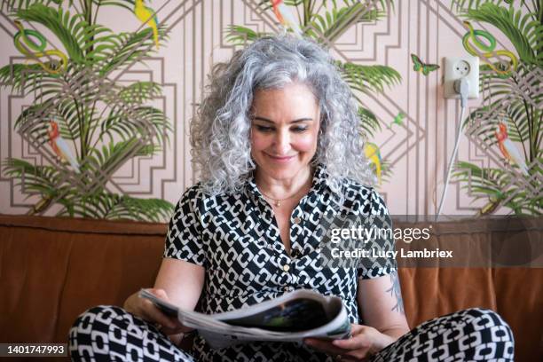 beautiful gray-haired woman reading a newspaper on the couch - vrouw behangen stockfoto's en -beelden