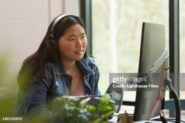 young asian woman works alone on computer - computer headset stock pictures, royalty-free photos & images