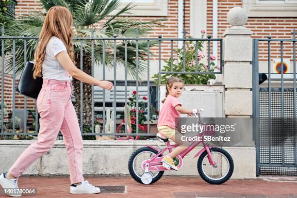 a woman watches her young daughter ride a bike with training wheels - stützrad stock-fotos und bilder
