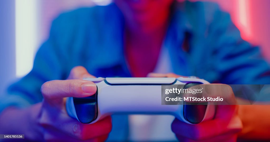 Nahaufnahme einer jungen asiatischen Frau, die eine Videospielkonsole im Neonlicht-Wohnzimmer zu Hause spielt.
