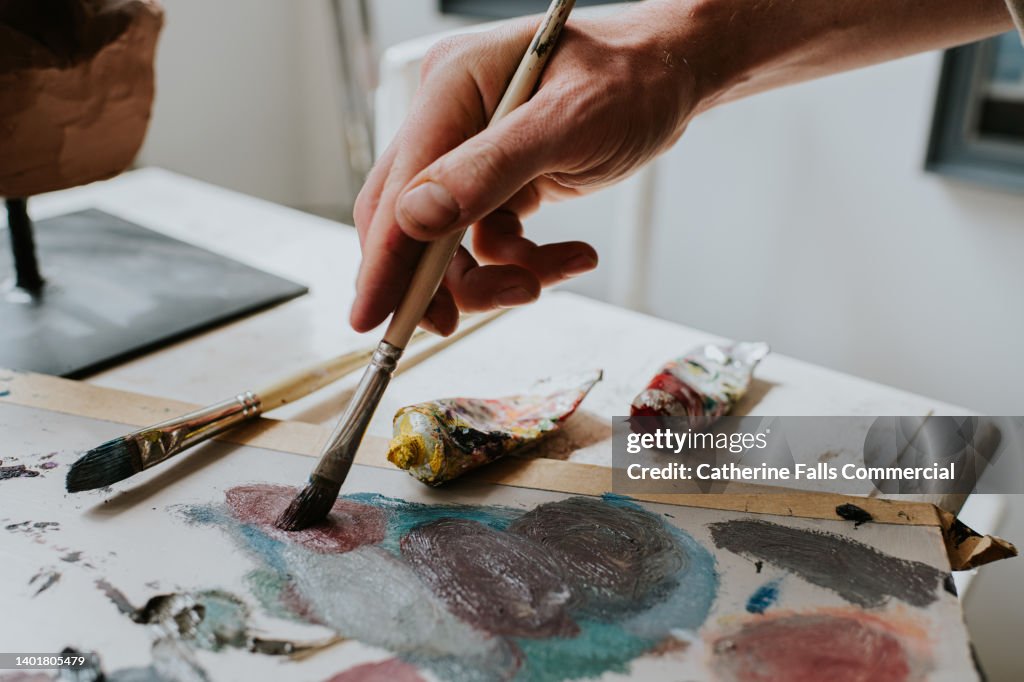 A hand holds a paint brush, and mixes oil paint on a board