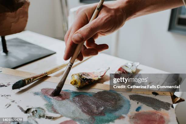 a hand holds a paint brush, and mixes oil paint on a board - painter artist fotografías e imágenes de stock