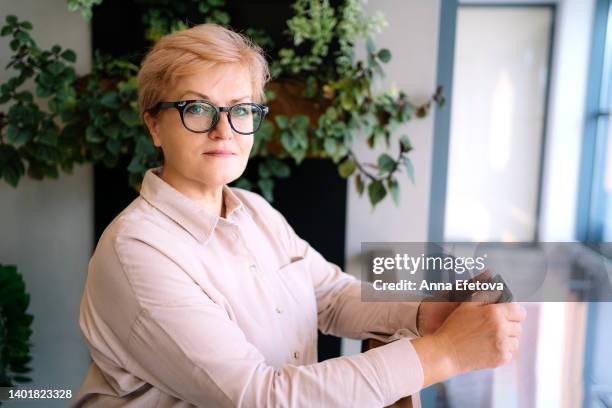 smiling senior blond haired woman with glasses stands against background of plants. she is wearing light colored blouse - grüne bluse stock-fotos und bilder