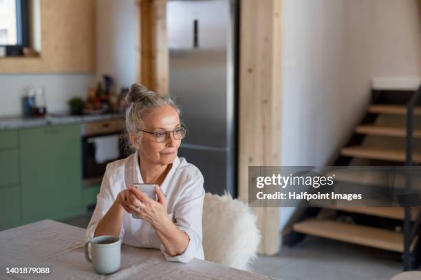 senior woman sitting in cottage kitchen with cup of tea, using smartphone. hygge atmosphere. - village home indoor stock-fotos und bilder