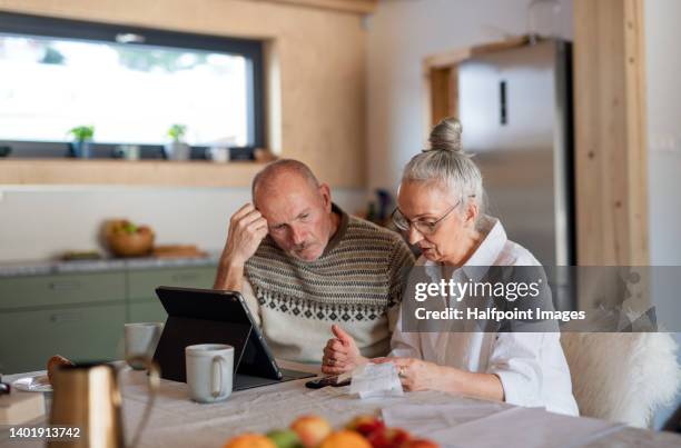 senior couple sitting at the kitchen table looking at digital tablet and recalculating their expenses. - physical pressure stockfoto's en -beelden