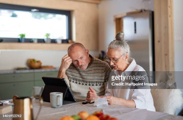 senior couple sitting at the kitchen table looking at digital tablet and recalculating their expenses. - consumerism stock pictures, royalty-free photos & images