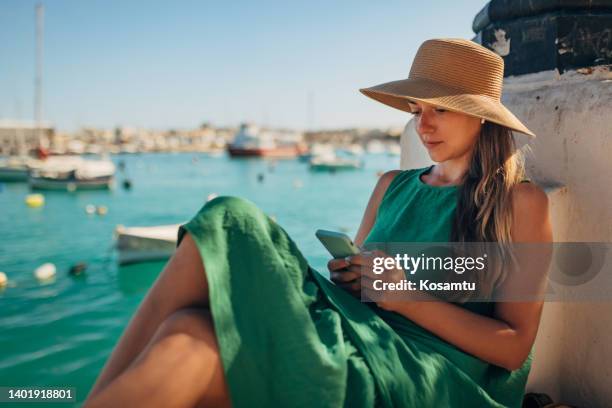 a millennial woman with wireless headphones sits near the sea and uses a mobile phone - malta boat stock pictures, royalty-free photos & images