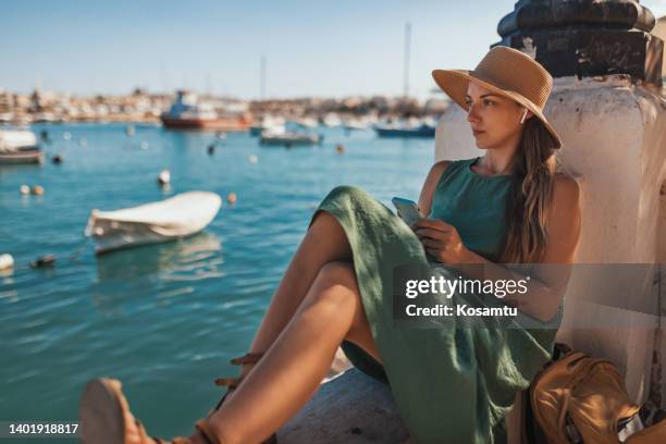 a beautiful woman in a summer dress and with a straw hat relaxing listening to music near the sea - sea iphone stock pictures, royalty-free photos & images